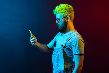 Profile of young adult bearded hipster guy with green hair wearing denim overalls, holding smart phone in hands, looks at display with serious expression. Colorful neon light, indoor studio shot.