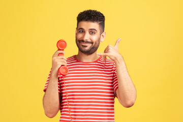 Positive bearded man in striped t-shirt holding red headset of retro landline telephone and showing...