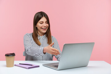 Happy smiling woman office worker welcoming client with handshake looking at laptop screen, talking on video call, online conference. Indoor studio shot isolated on pink background