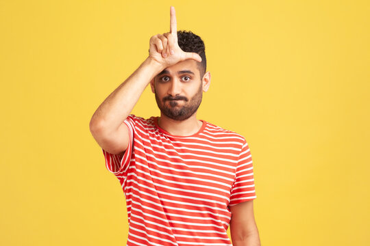 Gloomy depressed man with beard in striped t-shirt showing looser gesture holding fingers near forehead, sad because of silly mistake. Indoor studio shot isolated on yellow background