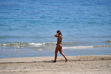Fototapeta premium A sportive young woman in a black bikini walks at the beach of Benidorm-Spain. A woman grabs her hair.