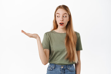 Image of excited girl looks at her hand holding copyspace with interest, display item on open palm, standing against white background. Advertisement concept