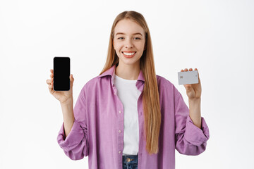 Portrait of smiling modern girl showing empty smartphone screen and credit card, introduce application, shopping discounts in online store, standing over white background. Advertisement concept