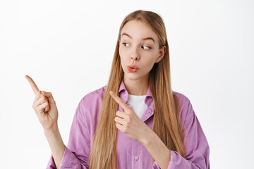 Beautiful blond girl looking curious left, pointing fingers at something interesting aside, showing...
