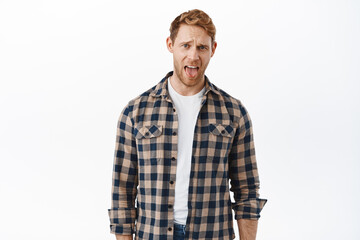 Handsome redhead man acting childish, adult ginger guy showing tongue and grimacing disgusted, hate bad taste, complaining, dislike something awful, standing over white background