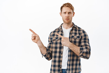 Sad and disappointed redhead man frowning, pointing fingers left with coy upset face, showing...
