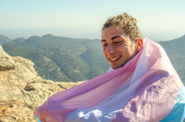Close up of Handome Transgender Gay Man Smiling Cute with LGTB trans flag blue and pink Covering him as a Cape at the Mountain Cliff on Sunny Day - obrazy, fototapety, plakaty