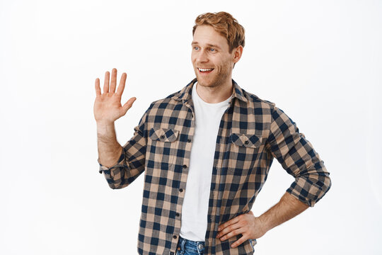 Image of friendly redhead man looking aside and waving hand to say hello, hey whats up, meeting a friend and greeting they with hi gesture, standing over white background