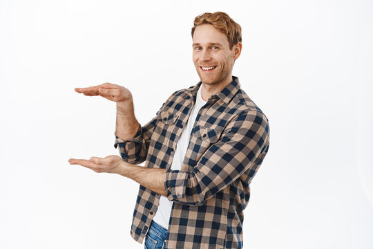 Image of strong and handsome redhead man holding your product logo against white copyspace, making empty box gesture and smiling at camera, display item between hands, white background