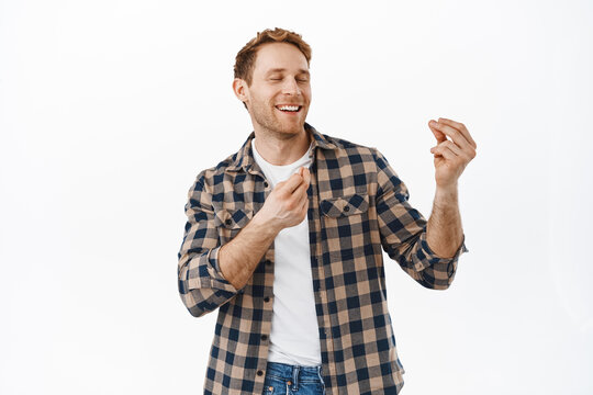Image of joyful redhead man snap fingers to rhythm of music, dancing with close eyes and satisfied face expression, enjoying party, celebrating success achievement, standing over white background