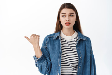 Skeptical, doubtful young woman looking and pointing aside at left copyspace with suspicious,...