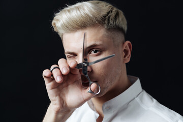 Portrait of blond male in white shirt stylist holding scissors near face over black background.