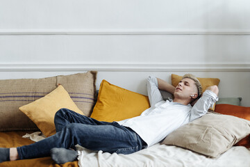 Blonde satisfied young caucasian man lying on comfortable sofa wear wireless earphones enjoy listen modern music audio book meditate relax feel no stress chill at home