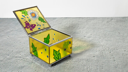 Open square old yellow glass jewelry box with painted flowers on a gray plastered background....