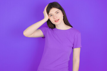 Portrait of confused young beautiful Caucasian woman wearing purple T-shirt over purple wall holding hand on hair and frowning, panicking, losing memory. Worried and anxious can not remember anything.