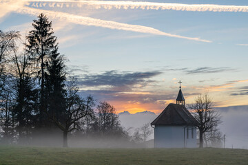 Fototapeta premium Kapelle im Winter bei Sonnenuntergang, im Hintergrund das Rheintal mit Nebelmeer und der Säntis im orangen Sonnenlicht. chapel in sunset with sea of fog and colored sky