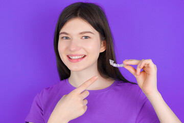 young beautiful Caucasian girl wearing purple T-shirt over purple background holding an invisible aligner and pointing at it. Dental healthcare and confidence concept.