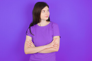 Pleased young beautiful Caucasian girl wearing purple T-shirt over purple background keeps hands crossed over chest looks happily aside