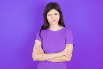 Fototapeta premium Gloomy dissatisfied young beautiful Caucasian girl wearing purple T-shirt over purple background looks with miserable expression at camera from under forehead, makes unhappy grimace