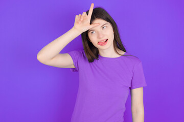 young beautiful Caucasian girl wearing purple T-shirt over purple background gestures with finger on forehead makes loser gesture makes fun of people shows tongue