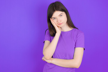 Sad lonely young beautiful Caucasian girl wearing purple T-shirt over purple background touches cheek with hand bites lower lip and gazes with displeasure. Bad emotions