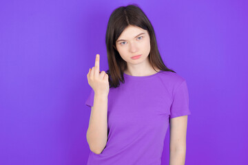 young beautiful Caucasian girl wearing purple T-shirt over purple background shows middle finger bad sign asks not to bother. Provocation and rude attitude.