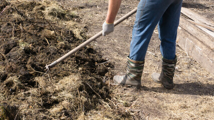 Farmer in blue trousers, rubber boots, and cotton gloves makes compost into the ground with a pitchfork with his own hands. Eco-friendly products