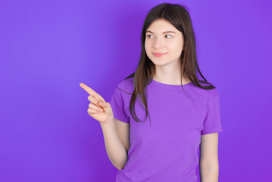 young beautiful Caucasian girl wearing purple T-shirt over purple background points at copy space and advertises something, advices best price.