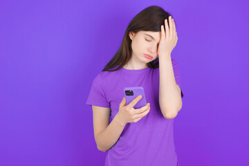 Upset depressed young beautiful Caucasian girl wearing purple T-shirt over purple background makes face palm as forgot about something important holds mobile phone expresses sorrow and regret blames