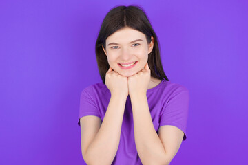 Satisfied young beautiful Caucasian girl wearing purple T-shirt over purple background touches chin with both hands, smiles pleasantly, rejoices good day with lover