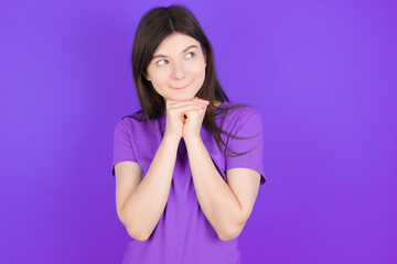 Curious young beautiful Caucasian girl wearing purple T-shirt over purple background keeps hands under chin bites lips and looks with interest aside.