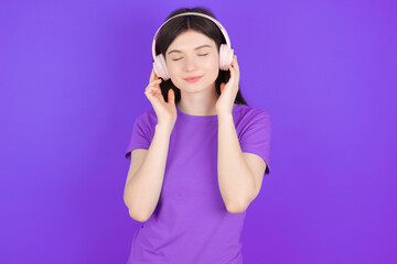 Pleased young beautiful Caucasian girl wearing purple T-shirt over purple background enjoys listening pleasant melody keeps hands on stereo headphones closes eyes. Spending free time with music