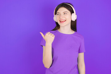 young beautiful Caucasian girl wearing purple T-shirt over purple background listens audio track via wireless headphones points thumb away advertises copy space