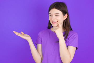 Positive young beautiful Caucasian girl wearing purple T-shirt over purple background advert promo touch finger teeth