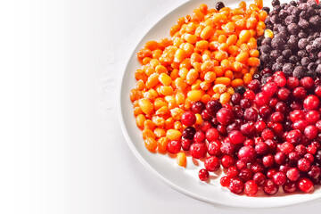 A horizontal composition with frozen sea buckthorn, cranberry and blueberry berries on a white plate on white background with copy space, close up. Storage, freezing berries