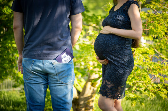 Junger, legere gekleideter Mann in Jeans und T-Shirt hat ein Ultrashallbild seines ungeborenen Kindes in der Hosentasche. Seine schwangere Frau steht vor ihm