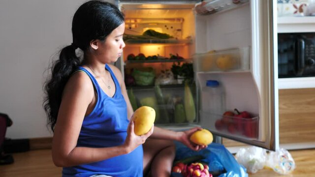A young pregnant woman organizing fruits and vegetables in the refrigerator.
