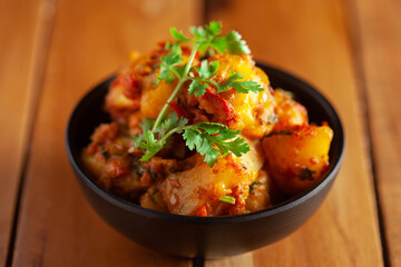 Close-up of Indian vegetarian dish of spicy Potato and Tomato curry garnished with green coriander fresh leaves. Served in a black ceramic bowl over wooden background.