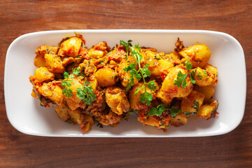 Close-up of Indian vegetarian dish of spicy Potato and Tomato curry garnished with green coriander fresh leaves. Served in a white ceramic plate over wooden background.