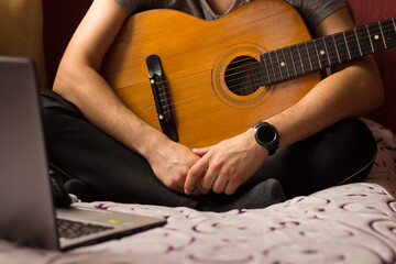 Fototapeta premium Learn to play the guitar online at home. A young guy with a guitar sits on the bed in front of a laptop and watches online guitar learning.