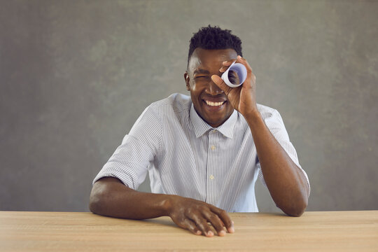 Studio portrait of happy curious black man looking with one eye through hole in paper roll. Cheerful positive African American guy in white shirt sitting at desk holding document scroll like telescope