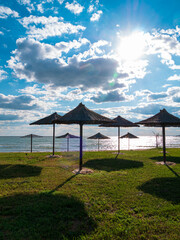 Straw umbrellas on green grass harsh shadow, sand beach vivid blue sea water sky background sunbeam. Scenic landscape wallpaper, summer vacation seasonal tropical marine travel rest resort advertising