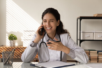 Glad to hear you feel well. Happy young latin lady doctor sit at desk talk to patient by phone...