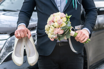 Groom dressed in blue suit holding a bridal bouquet of roses and white female shoes.