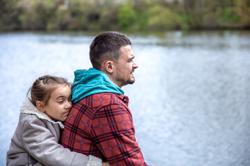 Fototapeta premium Little girl hugs dad, sitting on the background of the river.