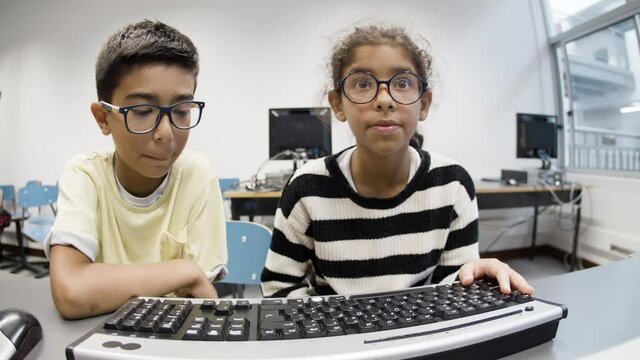 Schoolkids sitting at table and using computer. Funny boy in eyeglasses with excited girl learning computer science together. Digital education, friendship concept.
