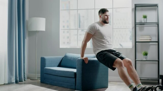 Skilled bearded trainer performs dip exercises on sofa edge