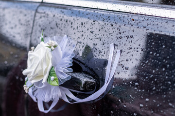 Faux rose boutonniere decorates a car door handle under rain.