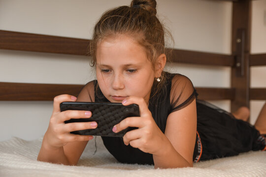 Technology, free time, communication concept. Cheerful teengaer dressed in nightwear, uses mobile phone for online chatting, networking, recieves text message, lies on bed