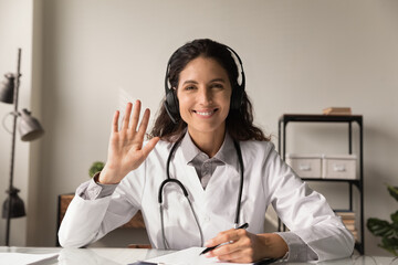 Telehealth. Headshot portrait of smiling latin woman doctor physician wear headphones look at camera wave hand to spectators. Young female medic consult remotely at virtual online meeting with patient - obrazy, fototapety, plakaty